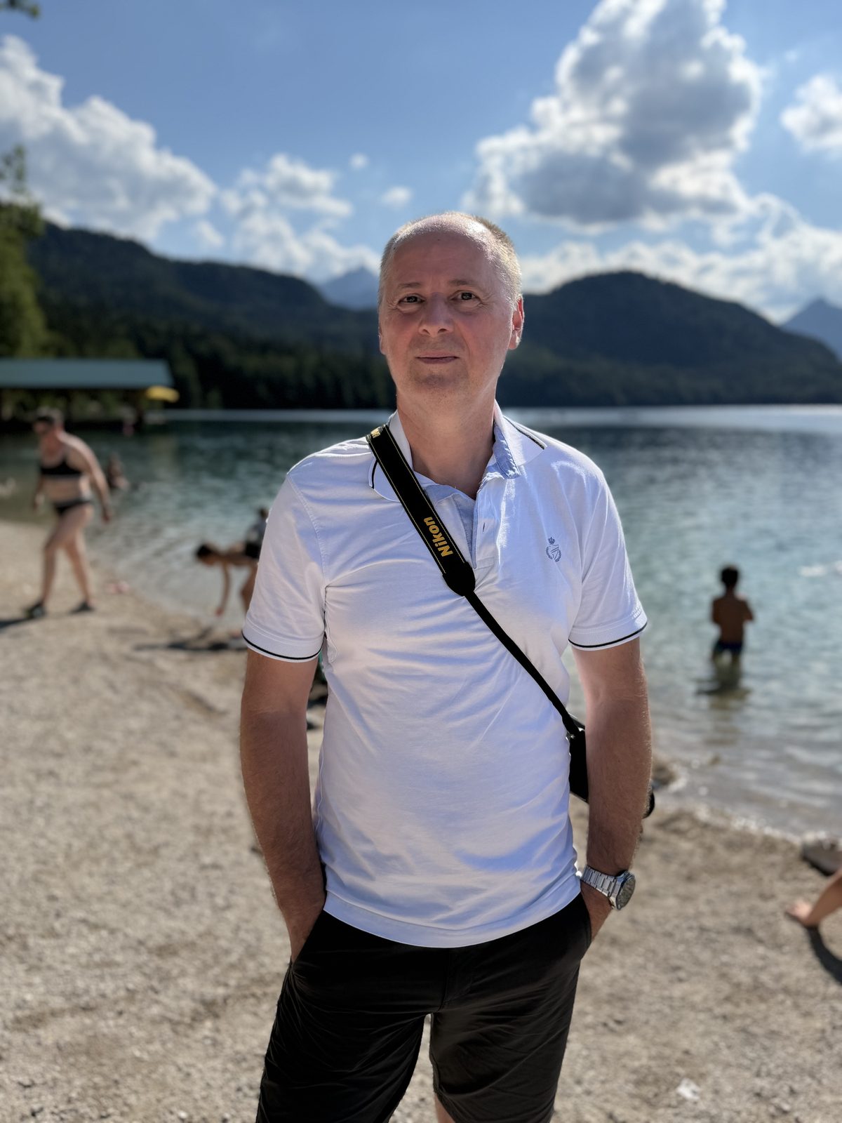 Férfi fehér pólóban fényképezőgép-szíjjal az Alpsee partján